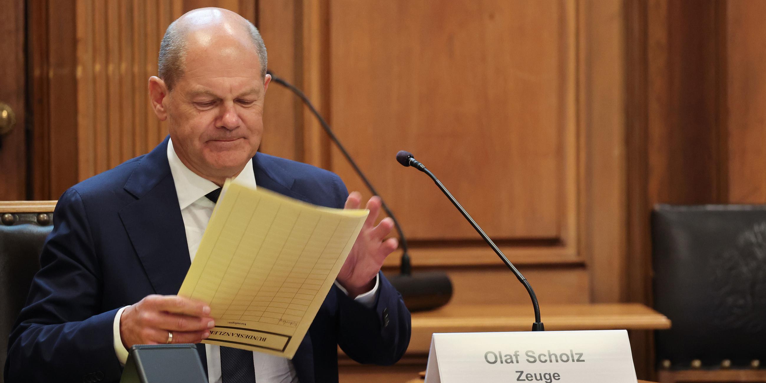 Bundeskanzler Olaf Scholz (SPD) sitzt auf seinem Platz auf der Senatsbank mit einem Plastikschild mit dem Text ·Olaf Scholz Zeuge· vor Beginn einer Sitzung des Parlamentarischen Untersuchungsausschusses ·Cum-Ex· im Plenarsaal der Hamburgischen Bürgerschaft im Rathaus. 