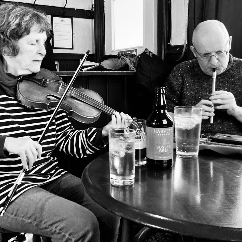 A woman playing fiddle and a man playing tin whistle at a small pub table.