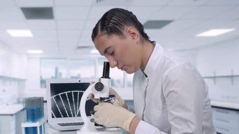 A female lab technician sitting at a table next to a laptop in a modern medical laboratory looks at biological samples under a microscope. A sample of DNA is being examined. 스톡 비디오