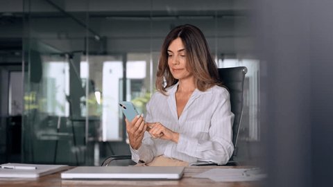 Smiling Latin Hispanic mature adult professional business woman using mobile phone cellphone. European businesswoman CEO holding smartphone using fintech application standing at workplace in office. - Βίντεο στοκ