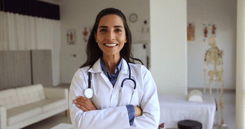 Happy Latina female general practitioner in white coat pose standing at workplace with arms crossed feels confident looks satisfied with career in private clinic. Professional medical worker portrait - Βίντεο στοκ