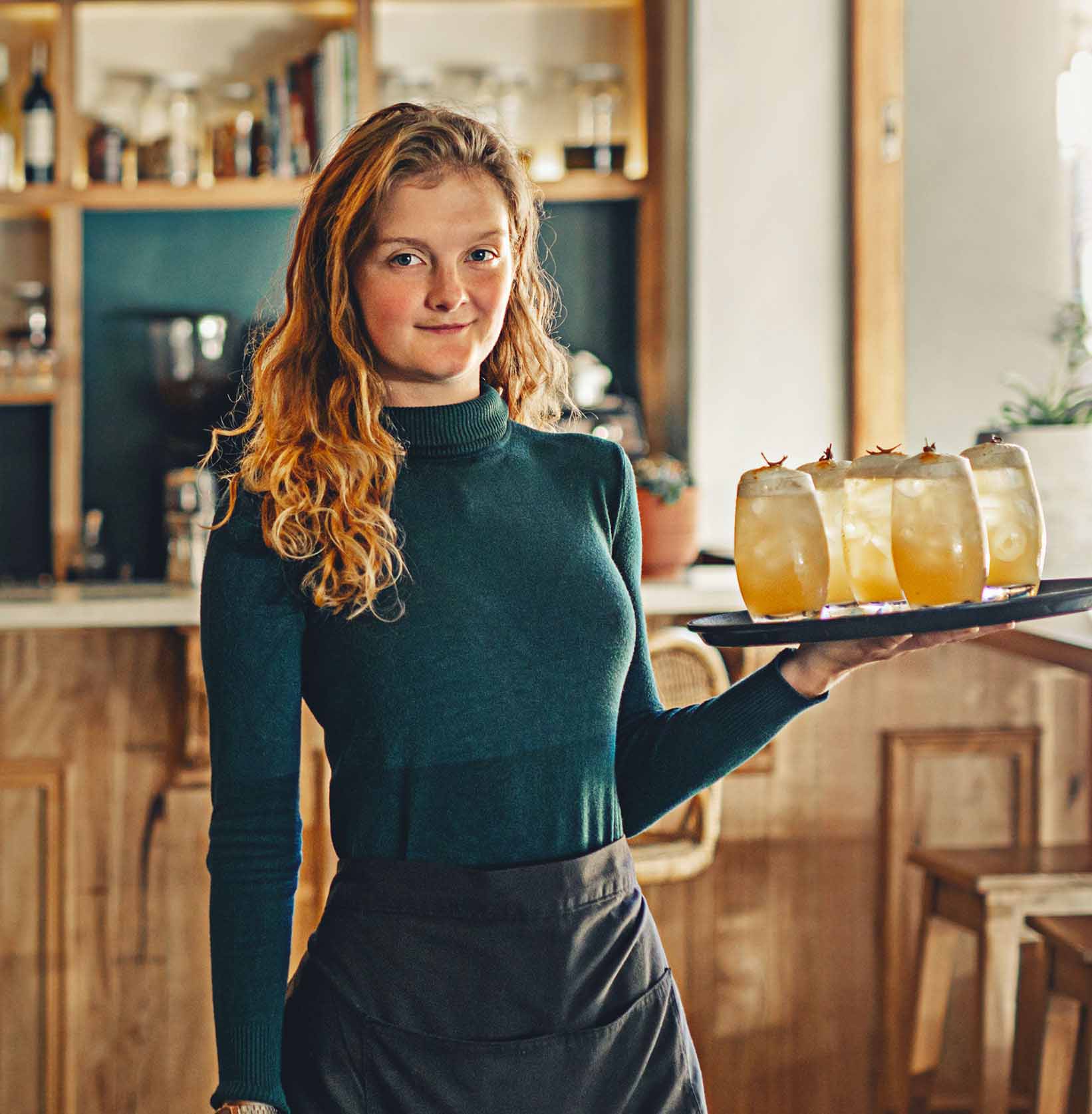 A woman smirking as she carries a tray with 4 drinks through the restaurant
