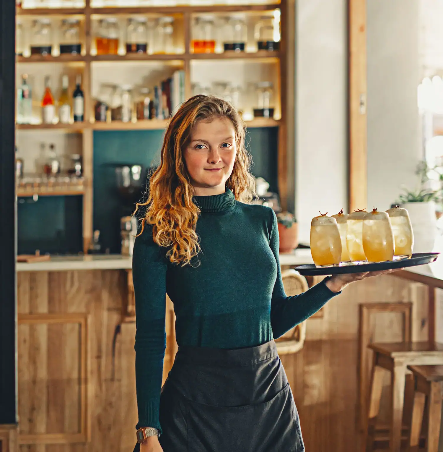 A woman smirking as she carries a tray with 4 drinks through the restaurant