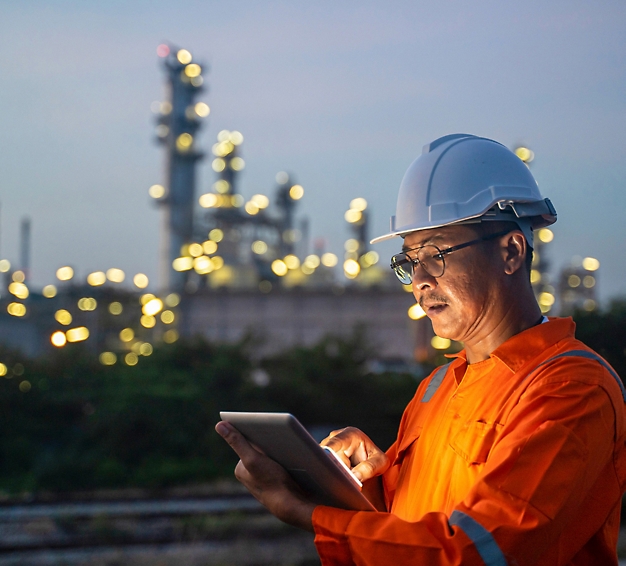 Engineer in orange overalls and hardhat using a tablet, with illuminated industrial plant in background at dusk.