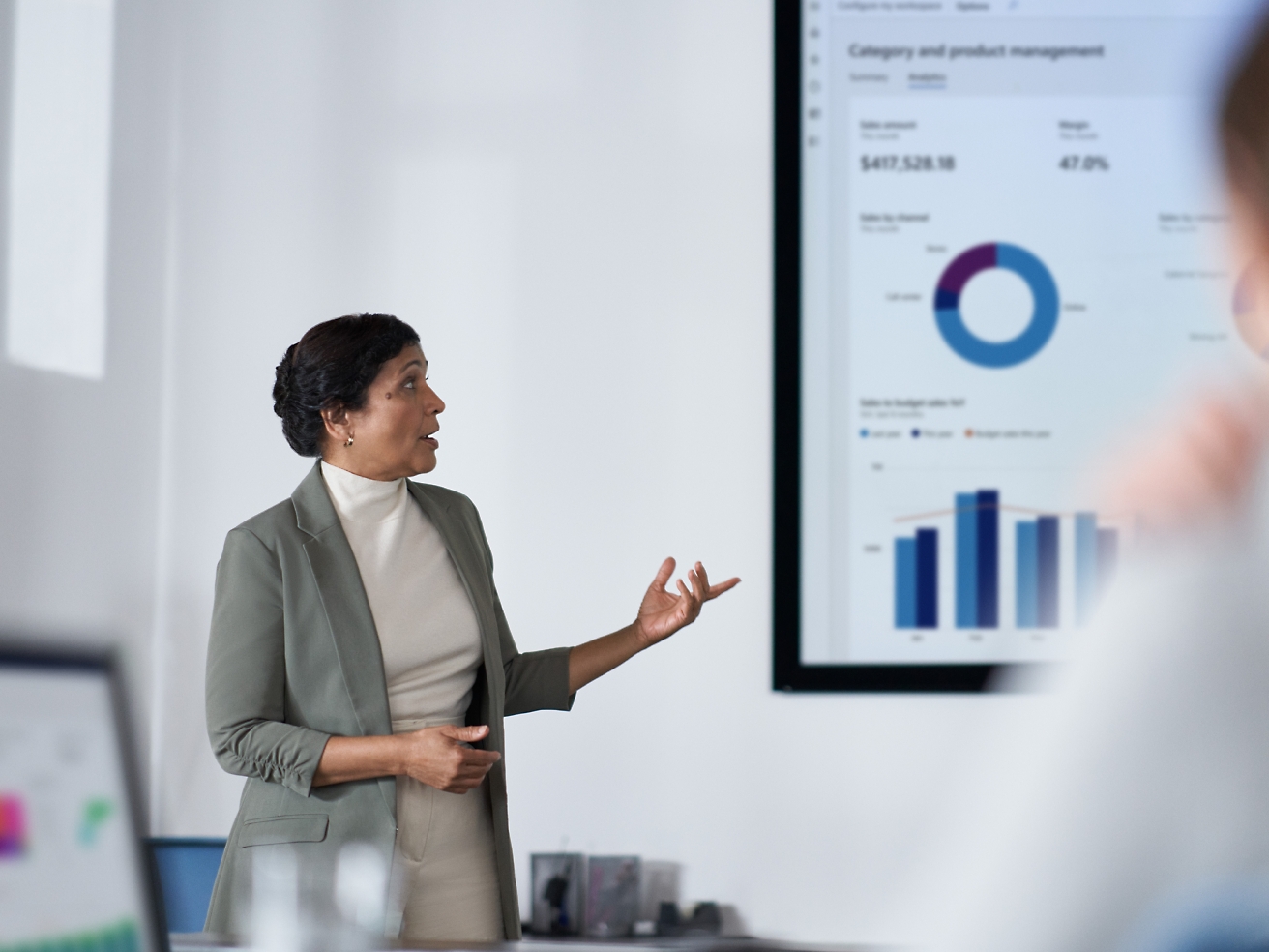 A professional woman presents financial data on a screen to her audience in a modern office setting.