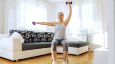 A smiling woman sits on a chair in her living room, doing a dumbbell workout. She's wearing a sports vest, three-quarter length leggings and sneakers. She holds two dumbbells; one of her arms is straight up overhead and the other is out to the side.