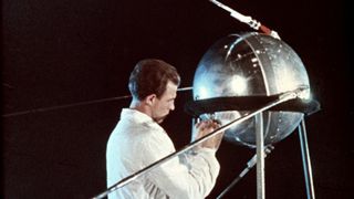 A person wearing a white laboratory coat works on a silver metallic satellite.