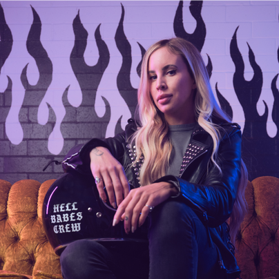 Jessica Wise, CEO of Hell Babes, sits confidently on a vintage sofa with a motorcycle helmet that reads “Hell Babes Crew” in front of a brick wall painted with black and purple flames