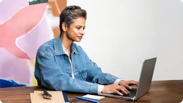 A woman with short hair wearing a blue denim shirt smiles while looking at her laptop on a wooden desk.