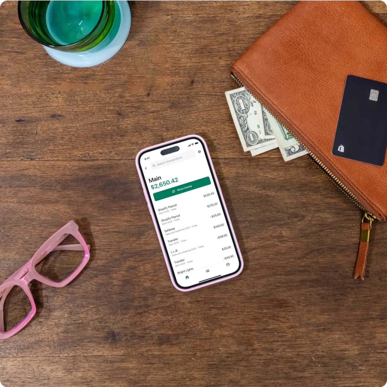 A mobile device on a wooden desk displays a Shopify Balance account balance, recent transactions, and a button that reads, “Move money.” Pink glasses, a wallet with US currency, and a green glass surround the phone on the desk.