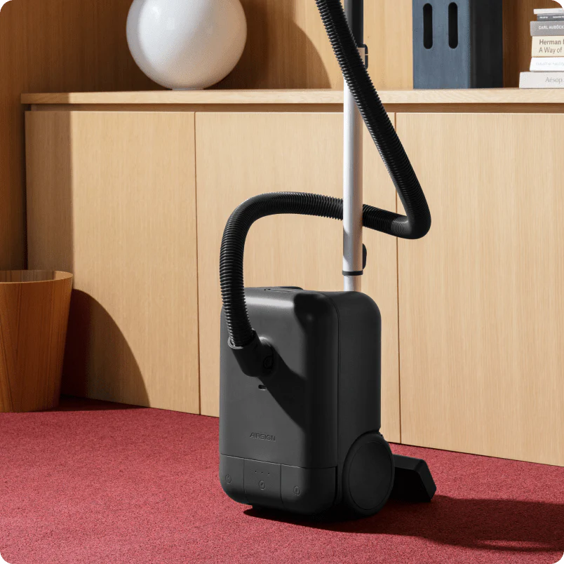 The sleek Airsign vacuum cleaner sits in a well-lit living room on red carpet, leaning against wood cabinets.