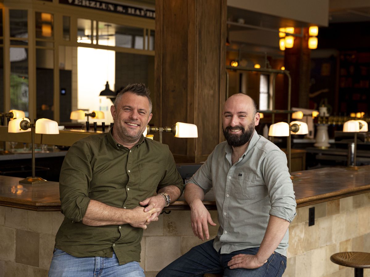 Two white men in collard shirts lean smiling against a stylish copper bar.