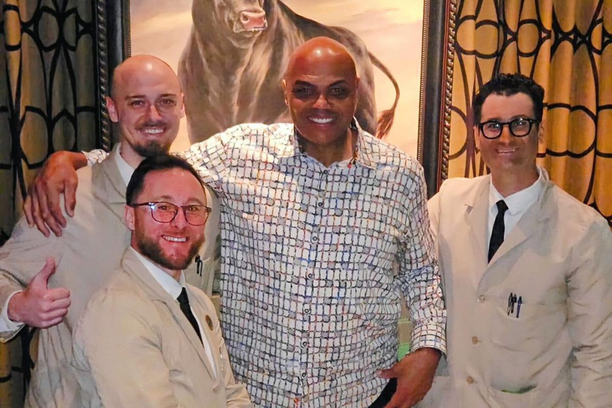 Charles Barkley in a blue and white patterned button-down shirt, standing with three serves from Manny’s, all dressed in a uniform of beige suit coats with white collared shirts and black ties. 