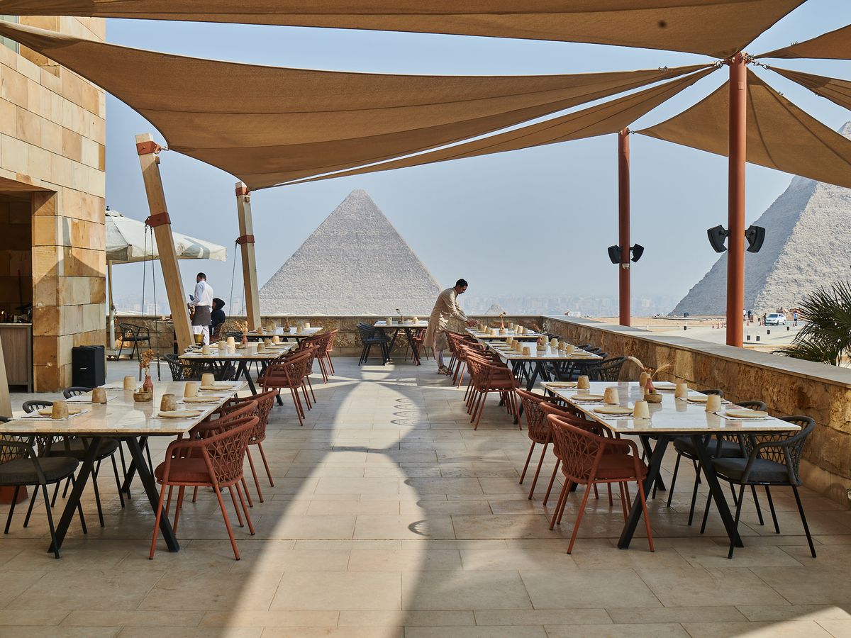 Diners and servers at outdoor tables on a patio, with the pyramids of Giza rising in the background. 