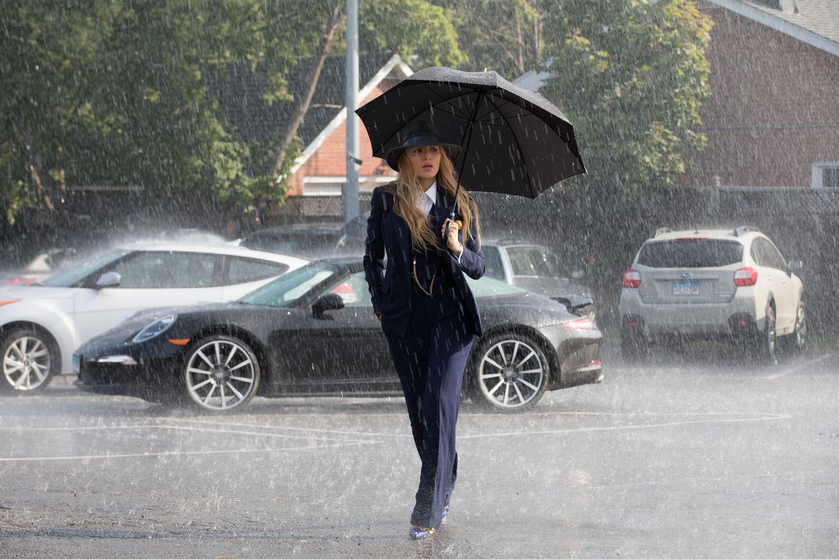 Blake Lively, wearing a dark suit and hat, walks in the rain with an umbrella in A Simple Favor