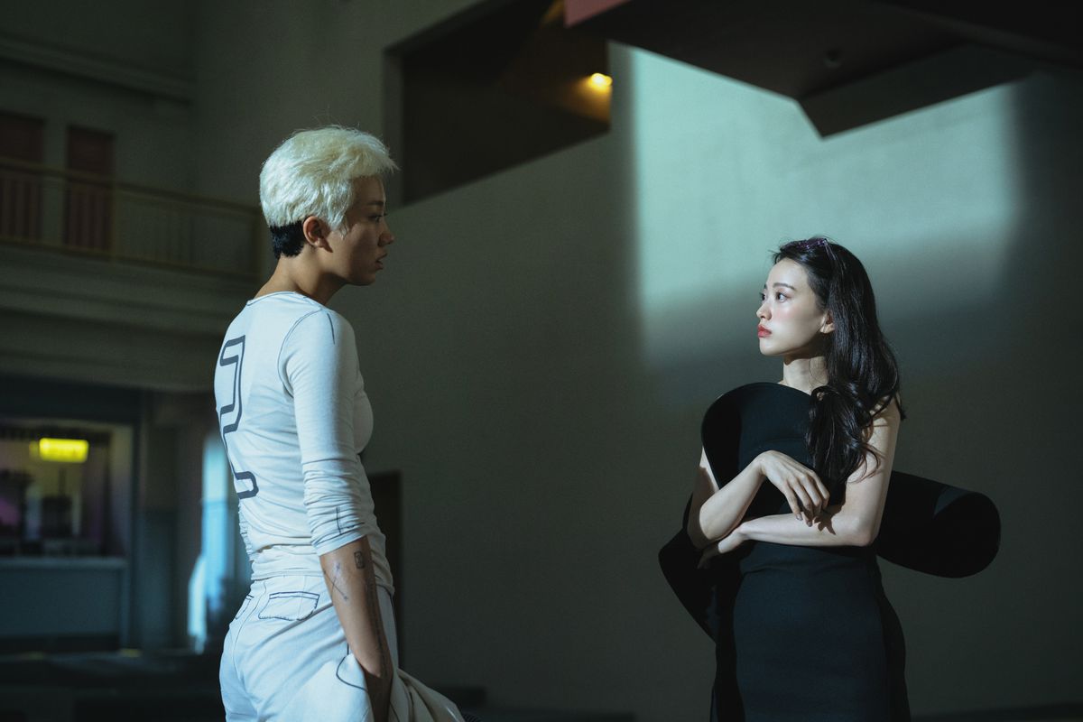 Two young people, one wearing a white uniform that has the number two on it, and the other wearing a black dress, talk in The 8 Show