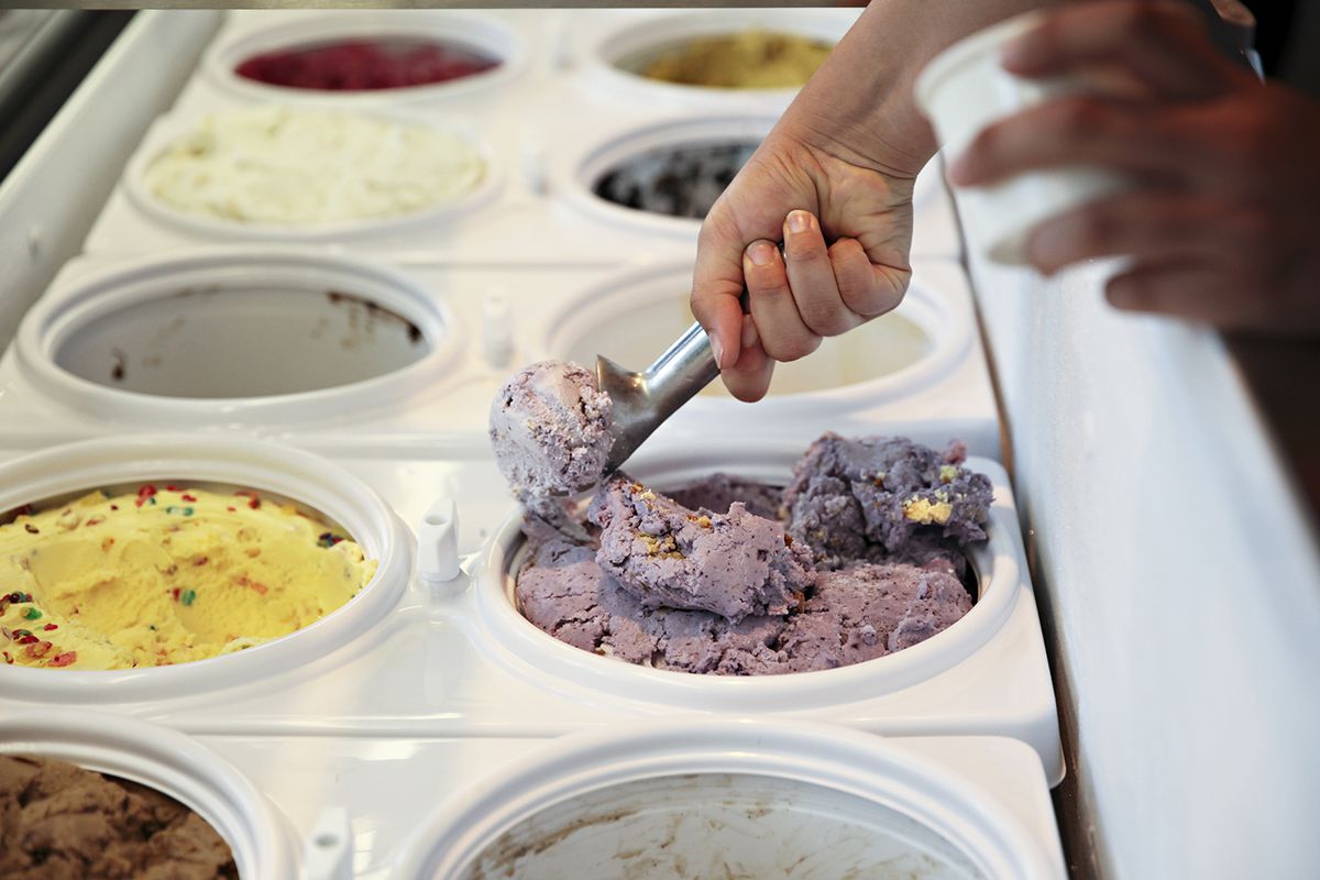 Two lines of ice cream buckets; a hand is scooping a purple berry flavor.