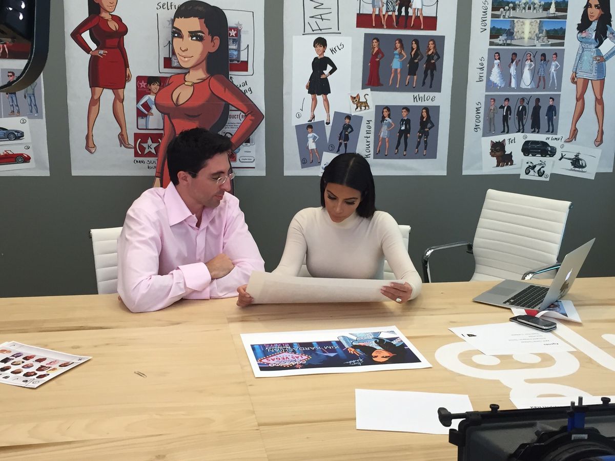 Glu Mobile CEO Niccolo de Masi, wearing a pink dress shirt, sits at a wooden table next to Kim Kardashian, wearing a cream-colored turtleneck, as they look over artwork from Kim Kardashian: Hollywood