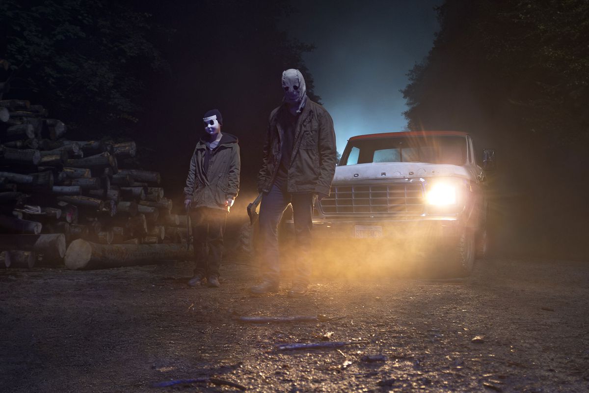 Masked strangers from The Strangers: Chapter 1 carry axes and walk away from a car with its headlights on. 