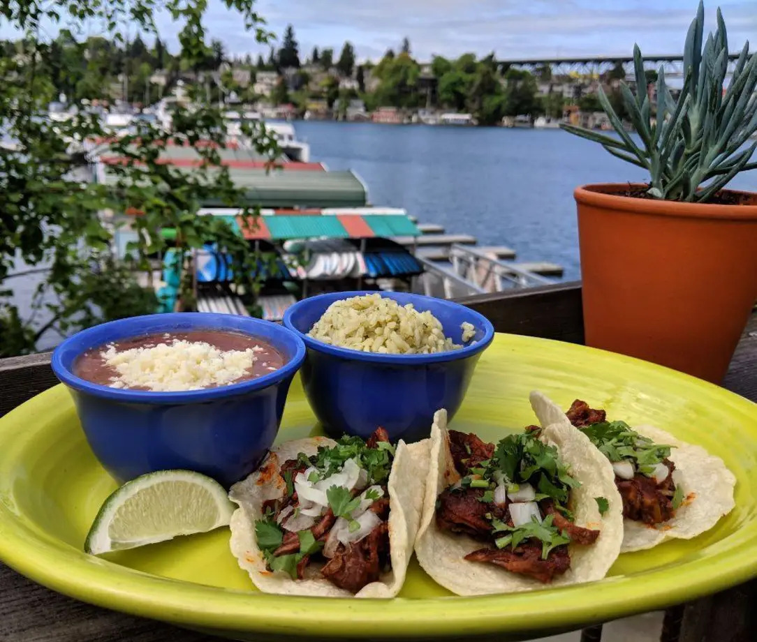 A yellow plate of pork tacos with blue cups of beans and guacamole, and a water view in the background.