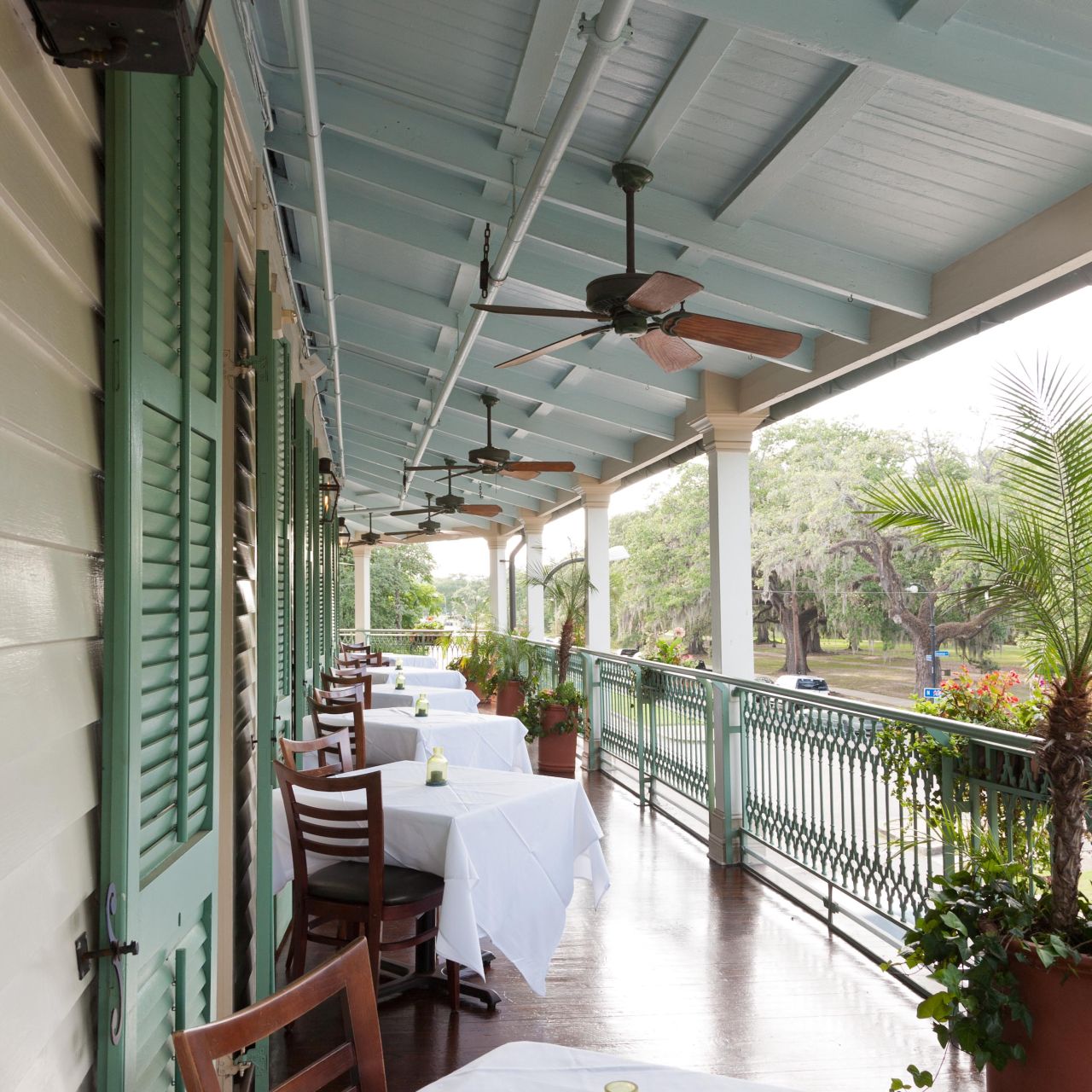 The second-story porch of a restaurant with small square tables set with white tablecloths, overlooking a park. 