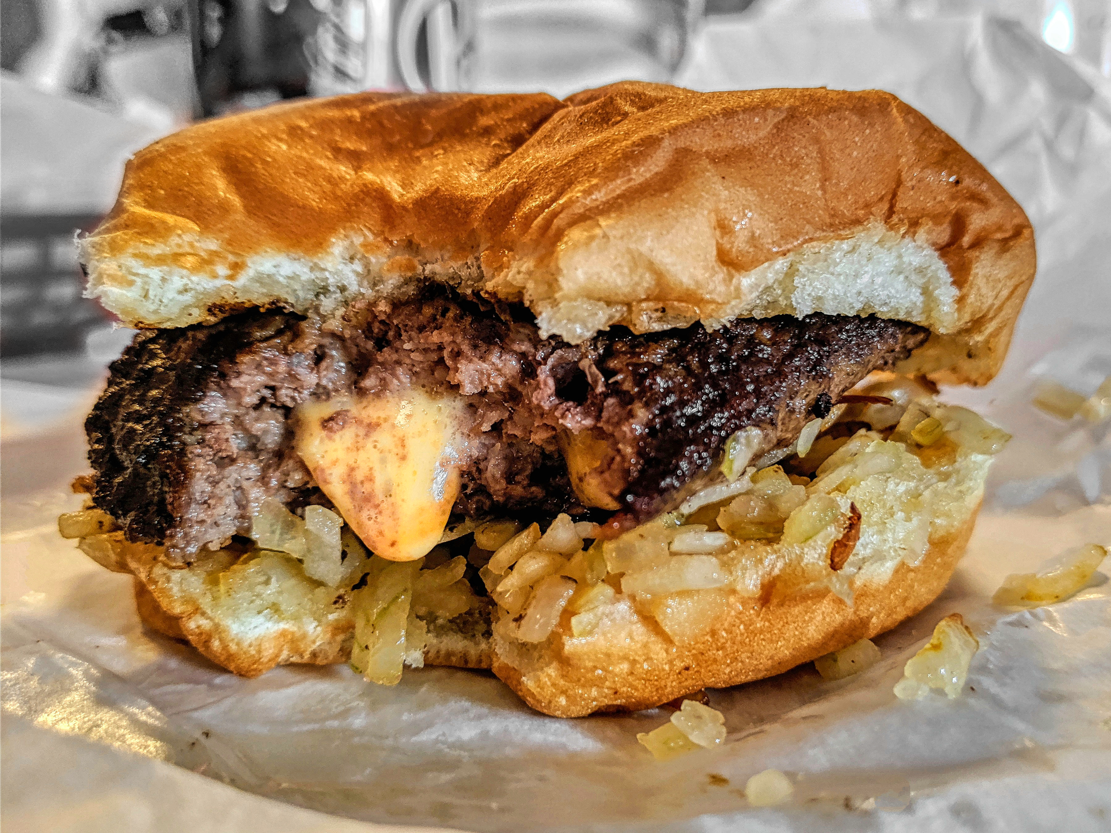 A Juicy Lucy burger on thin white paper, bitten into, with caramelized onions underneath the patty, yellow cheese oozing out of the meat, and a soft golden bun. 