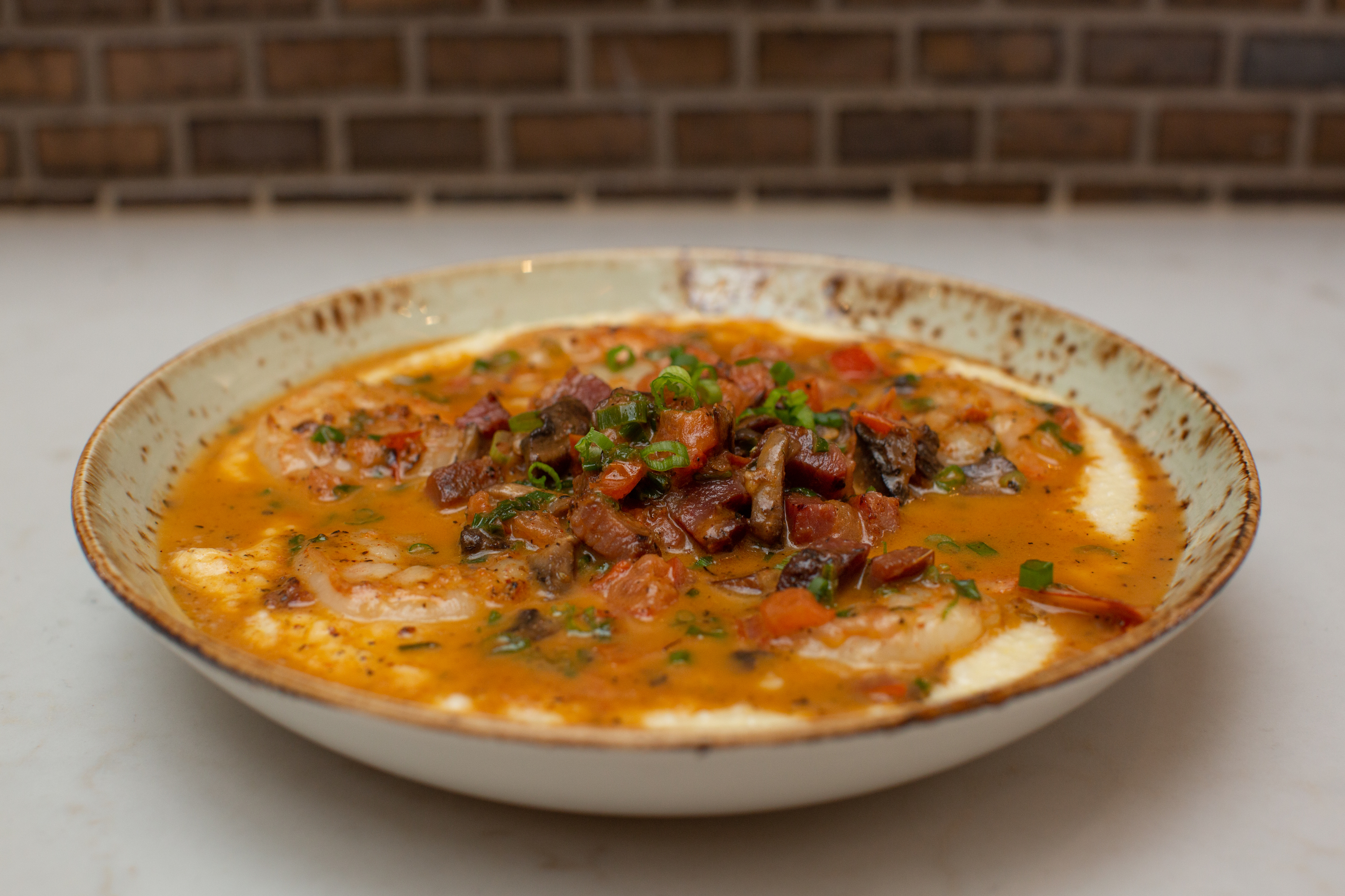 A bowl of creamy white grits dotted with large shrimp in a red-brown sauce and topped with pieces of sausage, tomato, and green onion.
