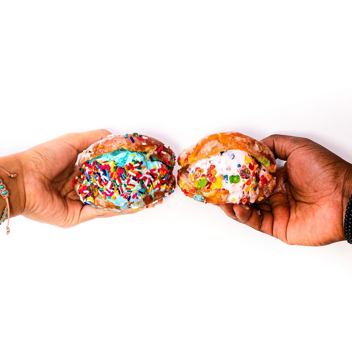 Two hands holding colorful doughnut sandwiches filled with ice cream. 