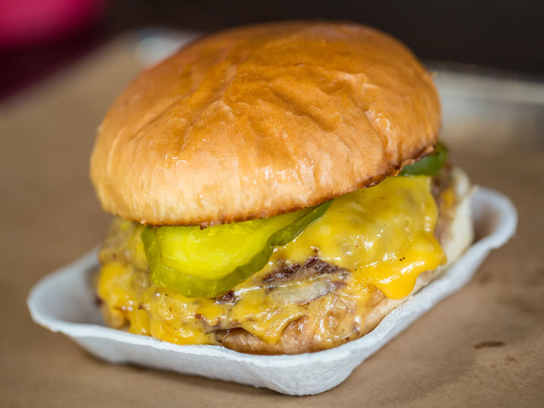 A cardboard plate holds a double patty burger covered in melted American cheese and topped with pickles on a plain, toasted bun. 