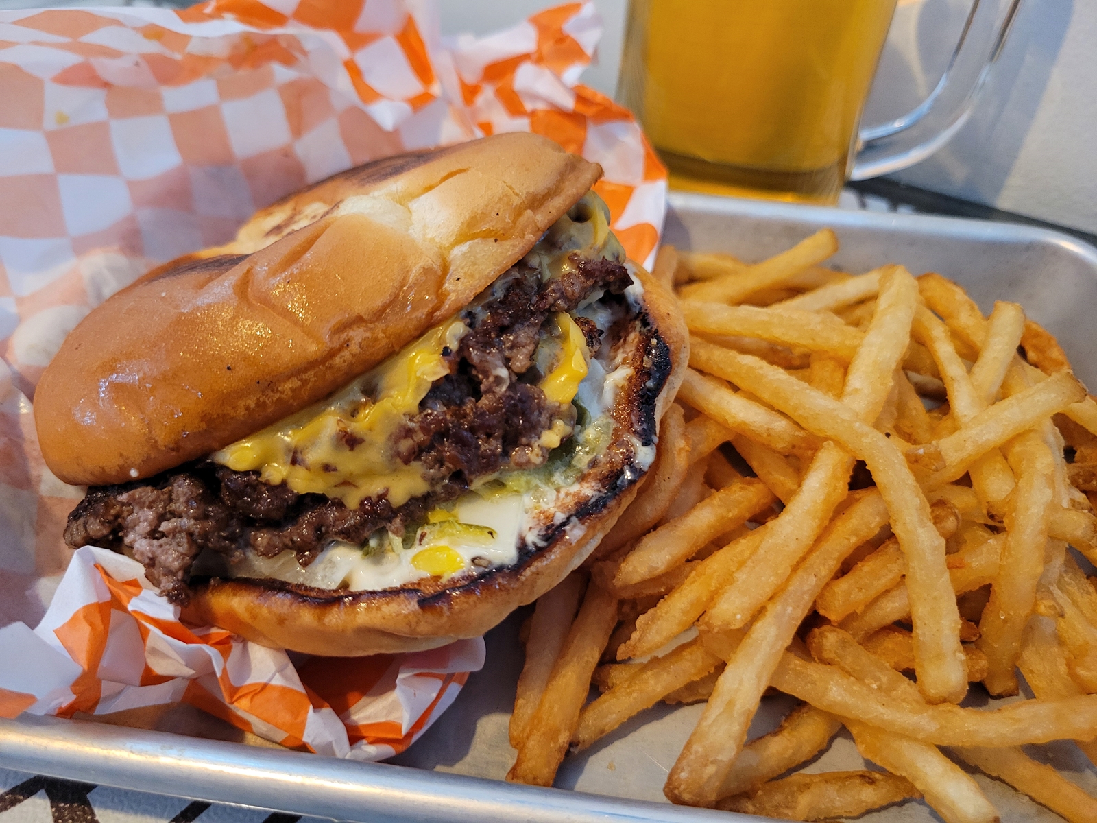 A burger in orange and white checkered paper with fries on a metal pan.
