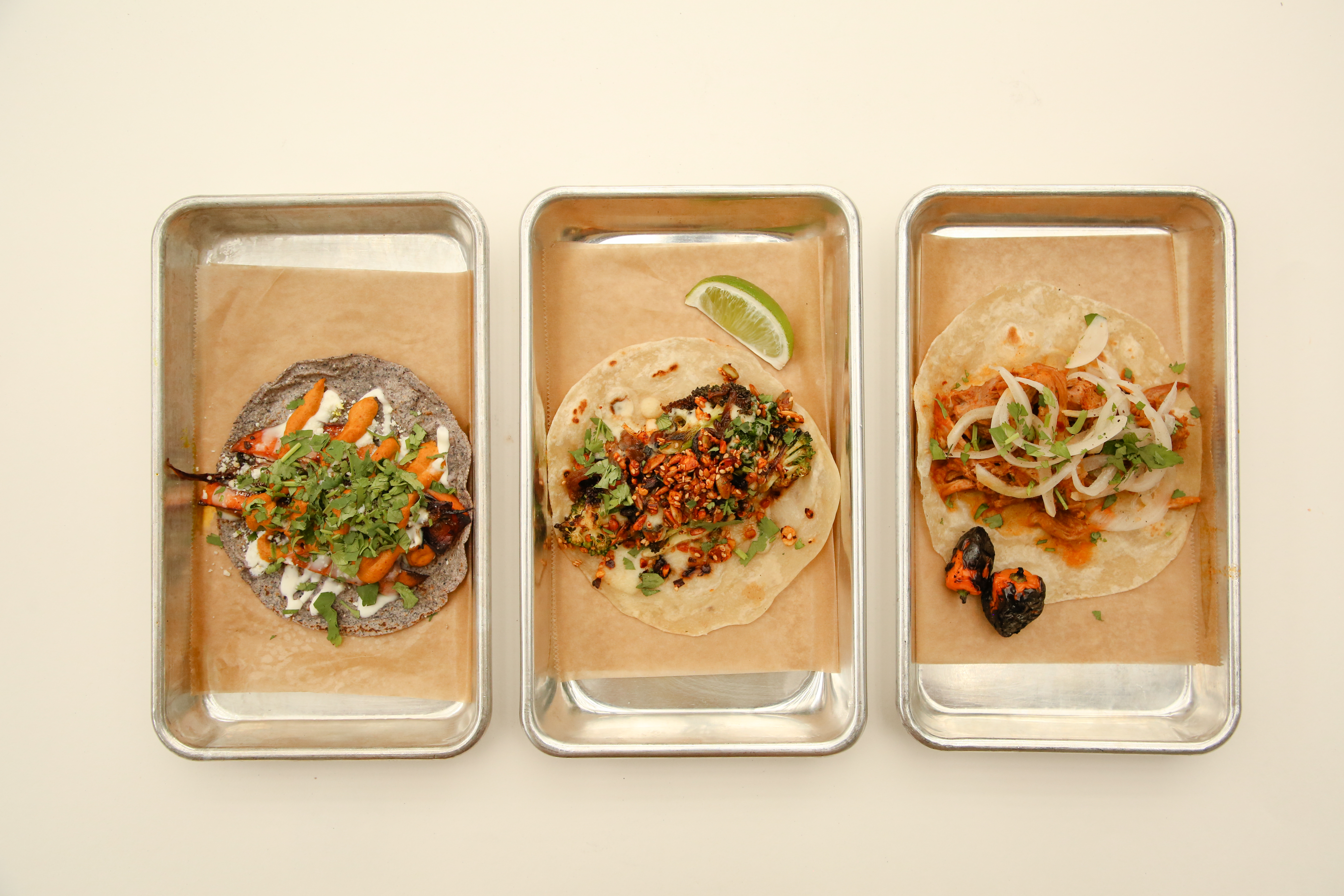 Three silver trays of tacos with brown paper underneath them on a beige background. 