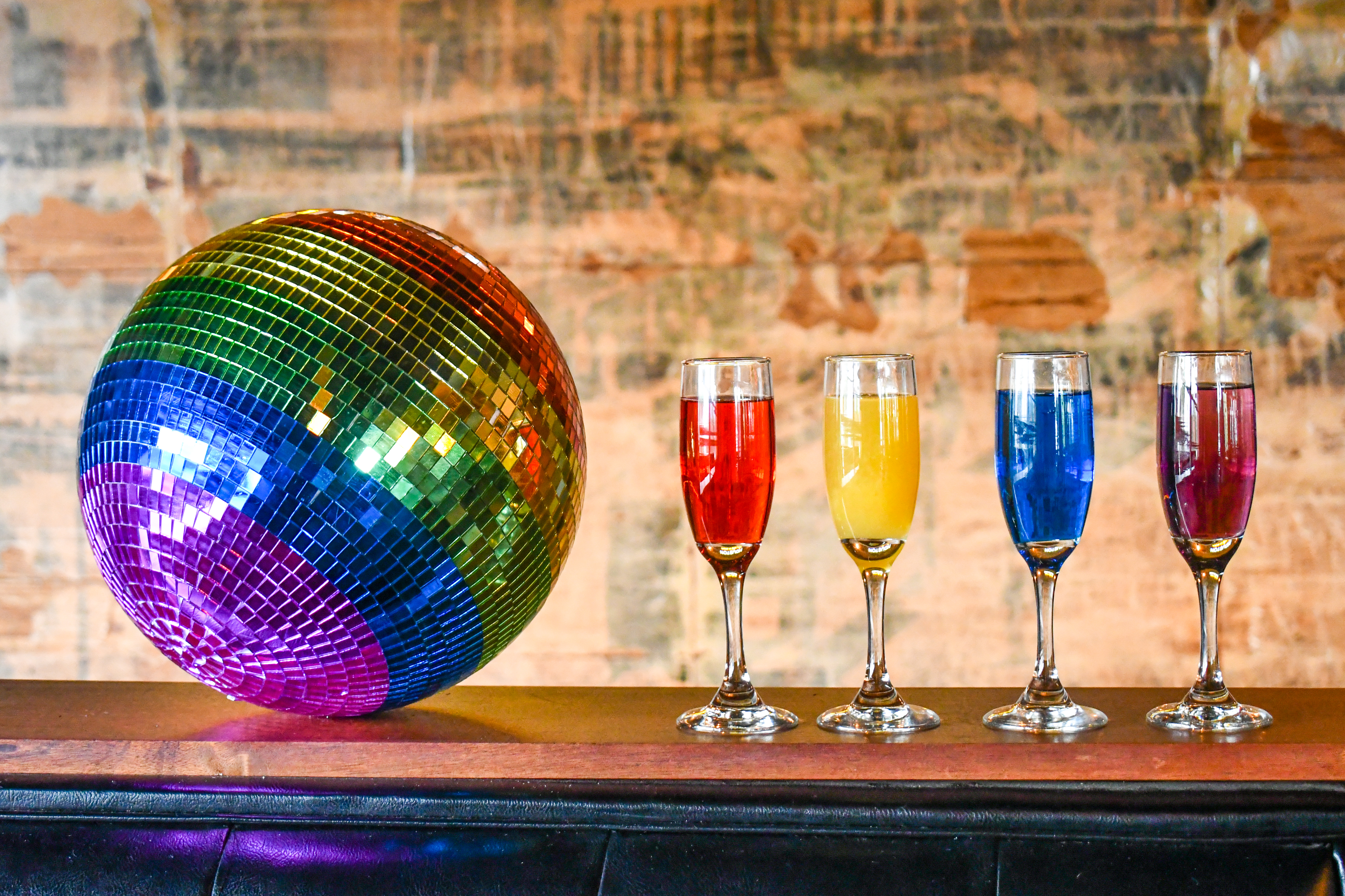 An image of rainbow-colored cocktails next to a rainbow-colored disco ball that’s sitting against a brick wall.