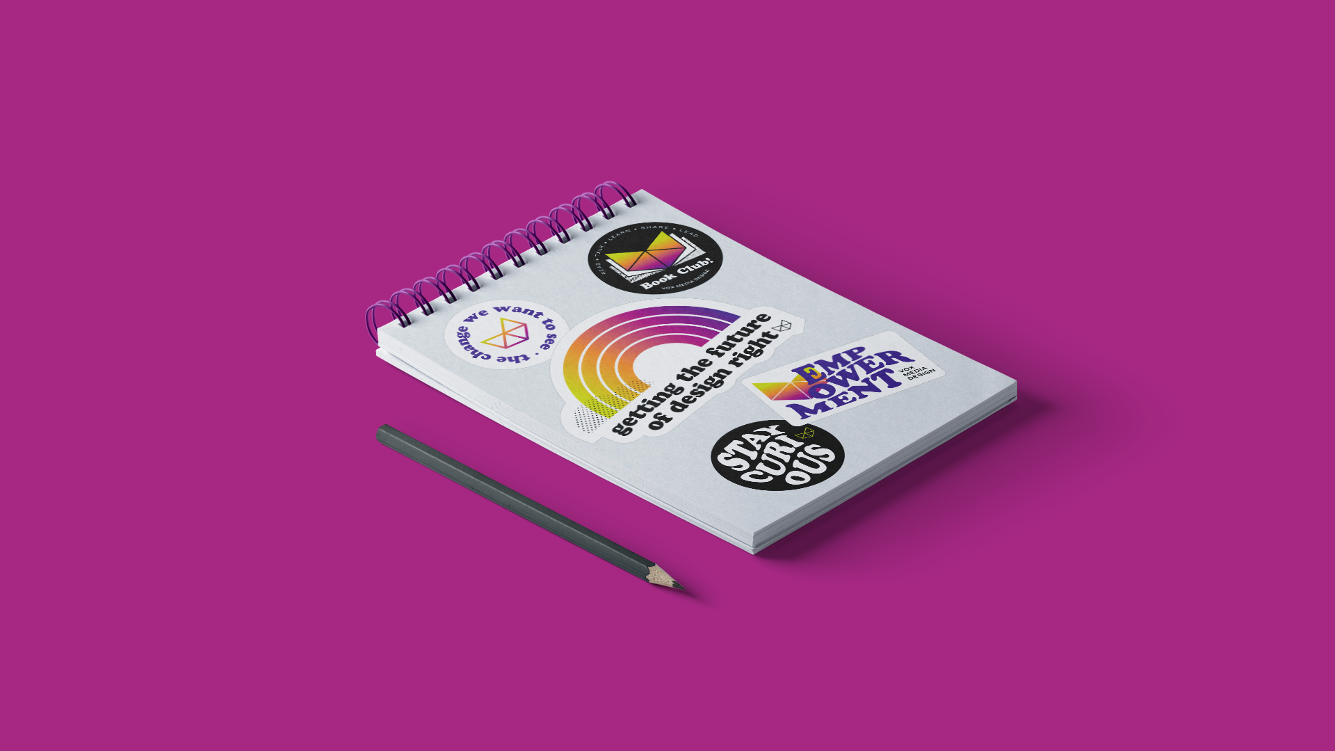 A notebook covered in Vox Media Design Team stickers, with a pencil, on a magenta desk