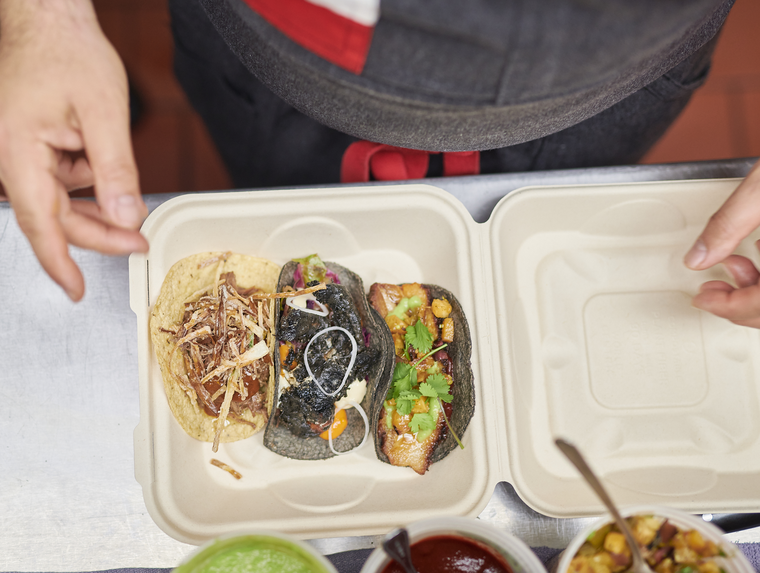 A brown paper tray with three tacos placed it it on a counter from an aerial view, with a person’s hands hovering over it. 