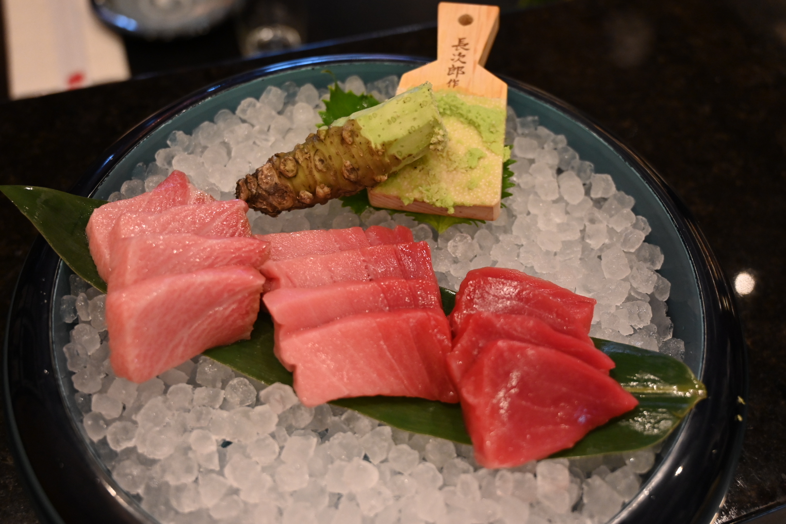Pink and red cuts of sashimi sitting on a green leaf on a plate of crushed ice, with a hunk of fresh wasabi root set beside them and a small piece of wood where it’s being ground. 