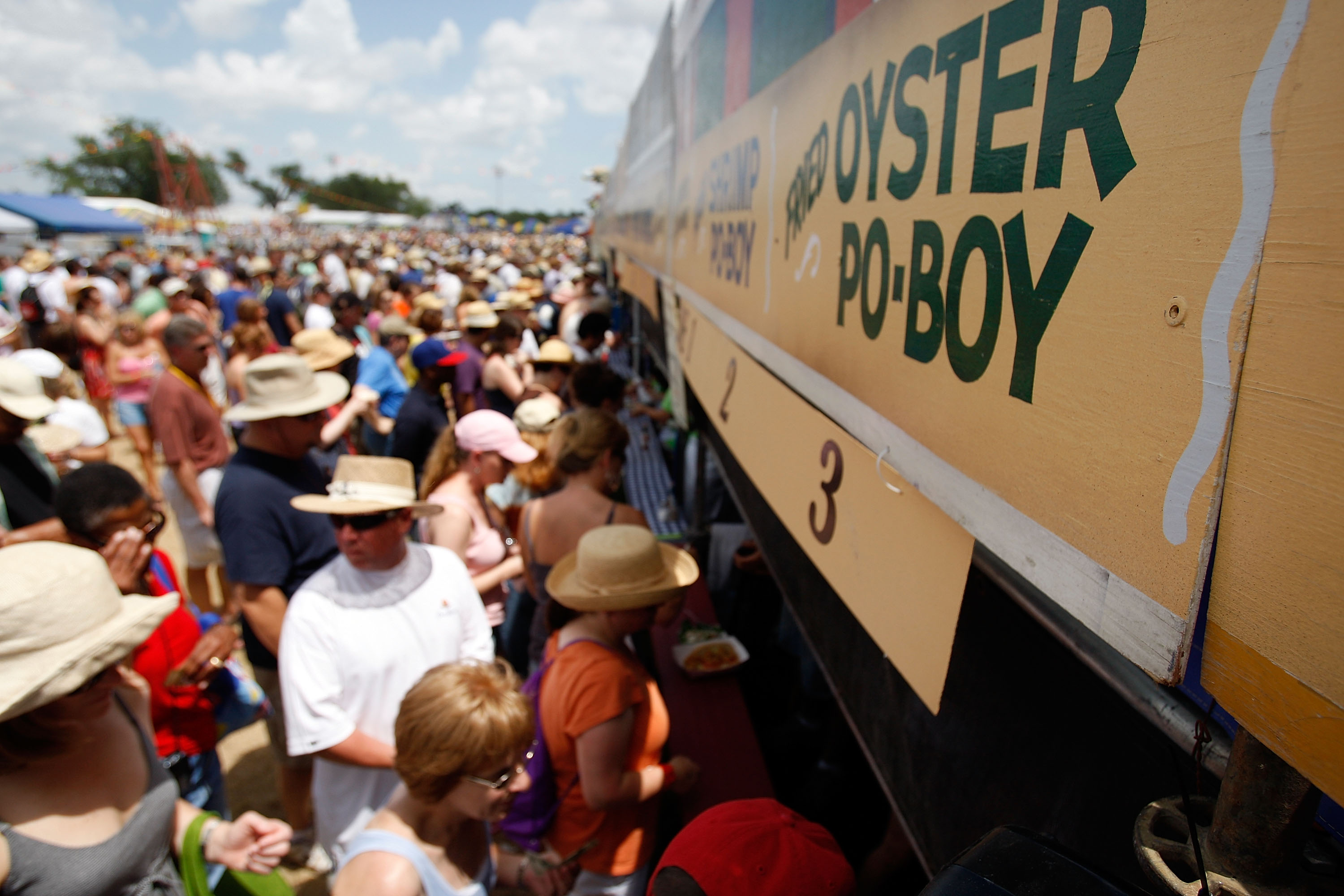 Crowds of people lining up to purchase food at Jazz Fest in New orleans, with a close-up view of the signage above the food stands visible in the foreground to the right. 