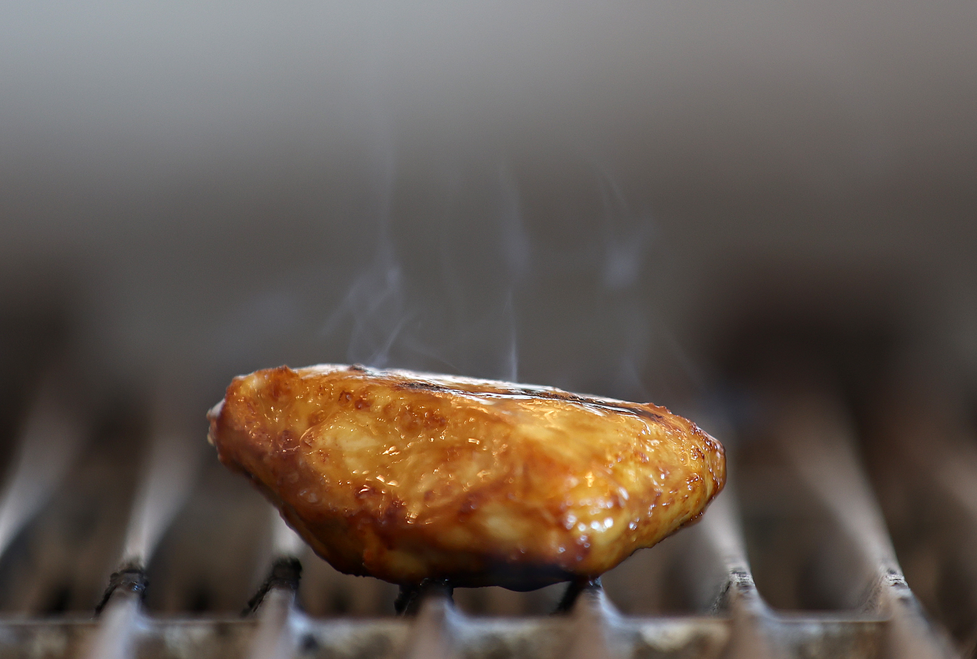 A piece of meat that looks like a chicken breast is covered in sauce and grill marks, steaming on top of a grill. 