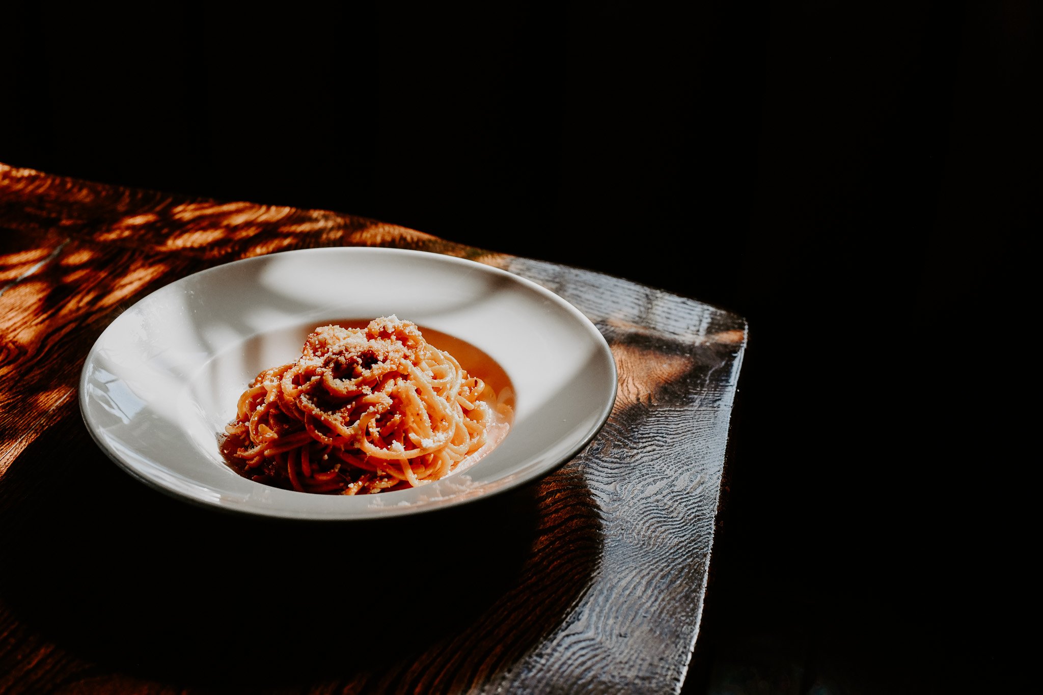 A white dish of spaghetti in red sauce sitting in a patch of sun on a table. 