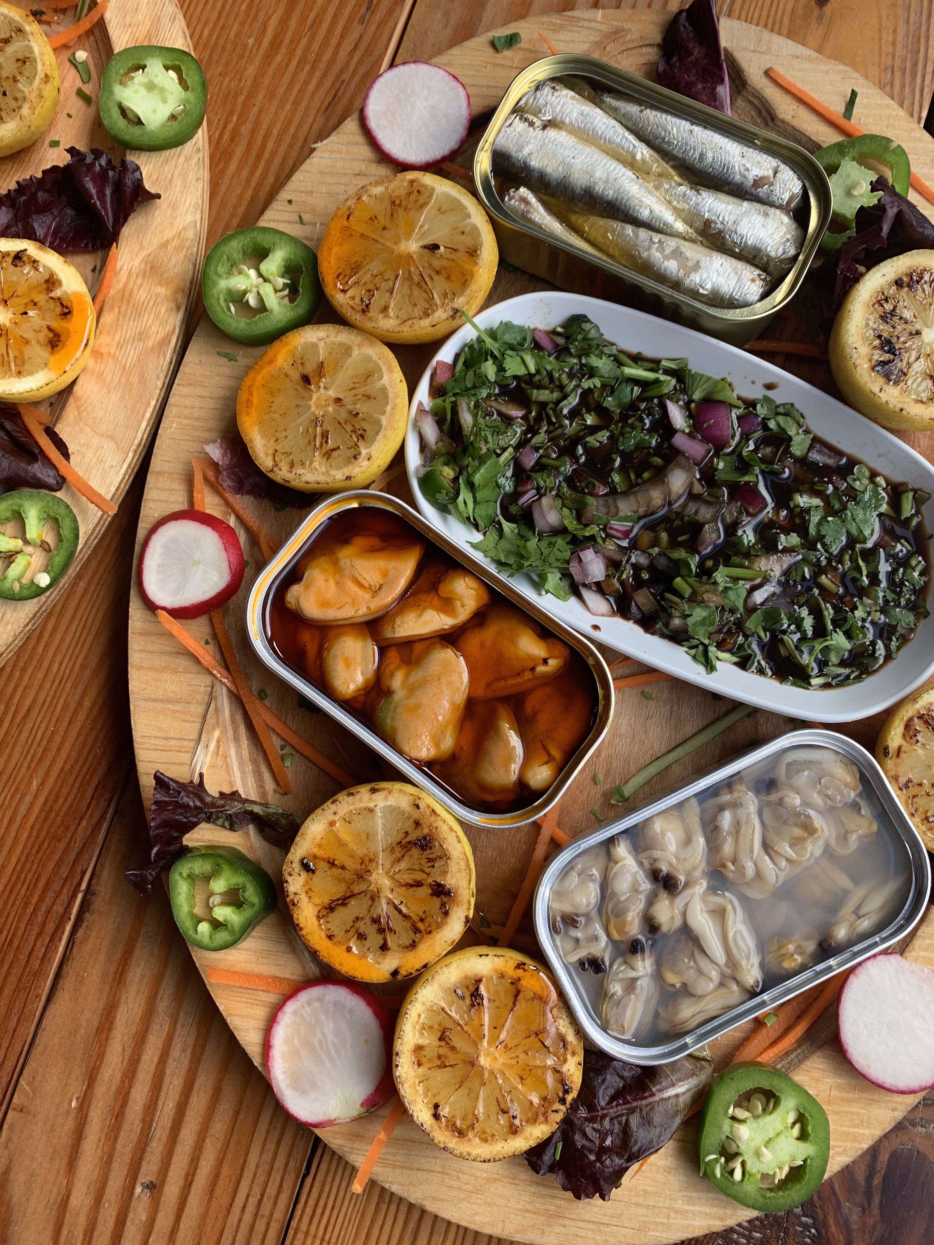 A platter of different opened tinned fish with charred lemons and cilantro garnish.