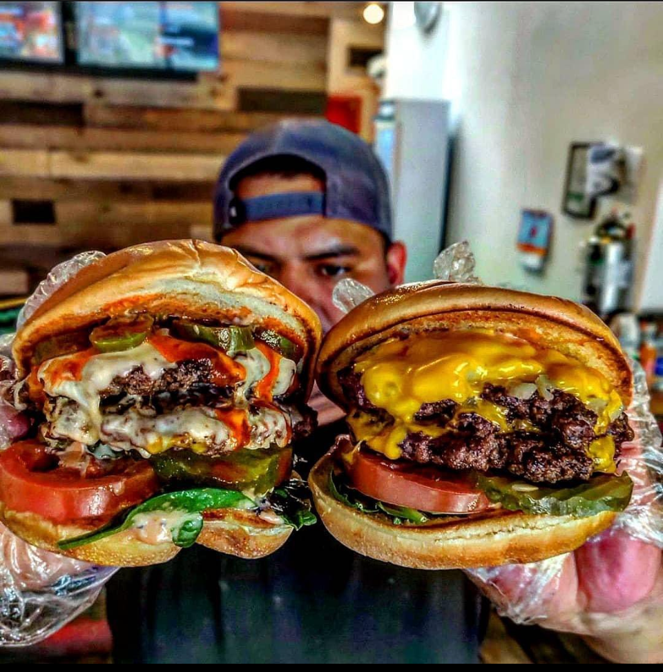 A person holding two burgers in each hand up close to the camera; the upper half of a person’s head wearing a baseball cap visible behind them.