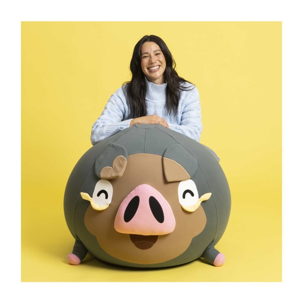 A photo of a person sitting behind a big round Lechonk beanbag. They’re smiling and there is a yellow background. The Lechonk looks really big and round.