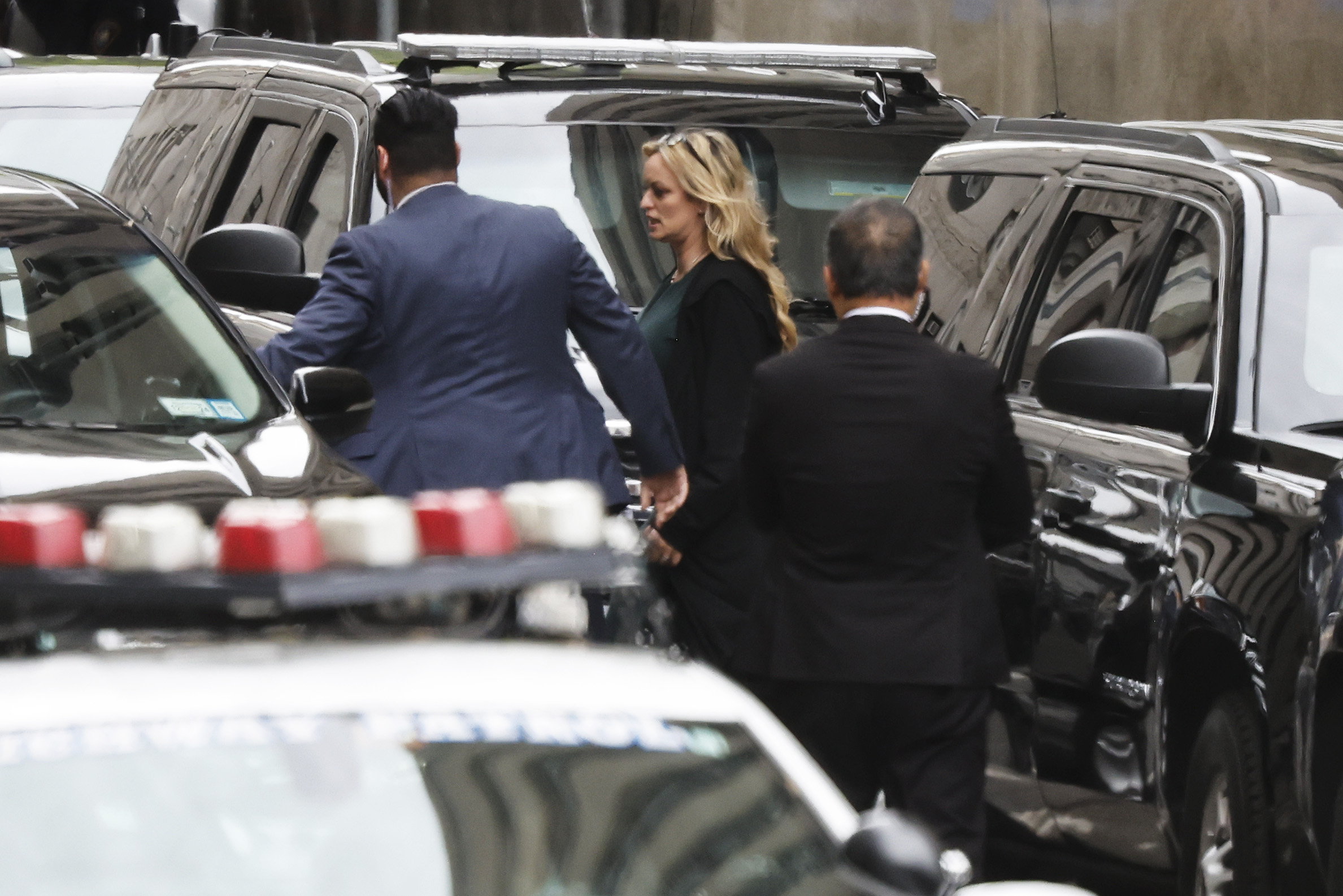 A blonde woman in black walking between parked SUVs with two men in suits.