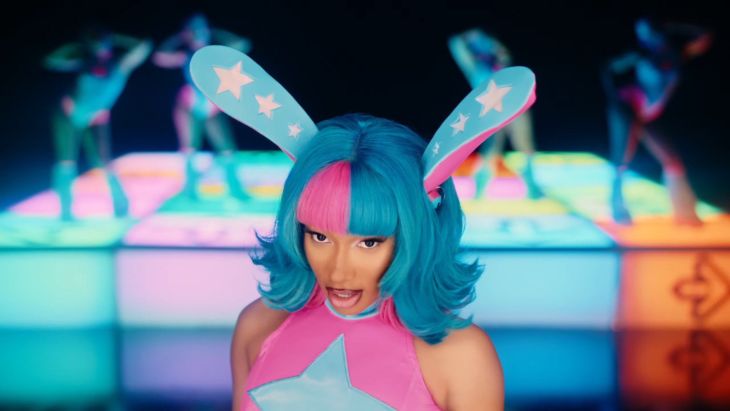 A still from Megan Thee Stallion’s video “BOA” in which the singer appears in a blue and pink anime rabbit girl outfit in front of a lighted Dance Dance Revolution-inspired stage with back-up dancers.