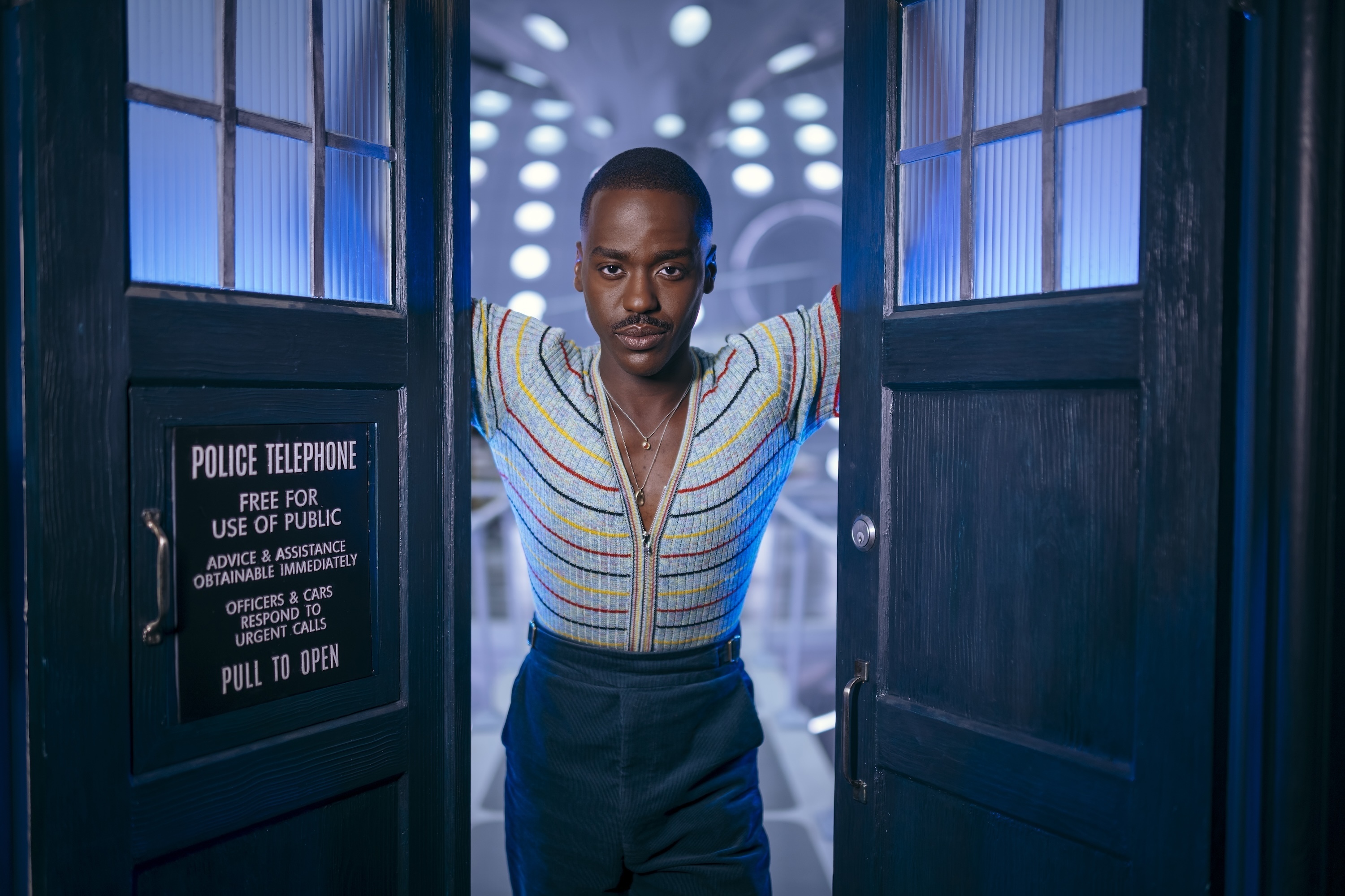 Ncuti Gatwa as the Fifteenth Doctor holding the twin doors of the TARDIS open as the ship behind him is visible. 