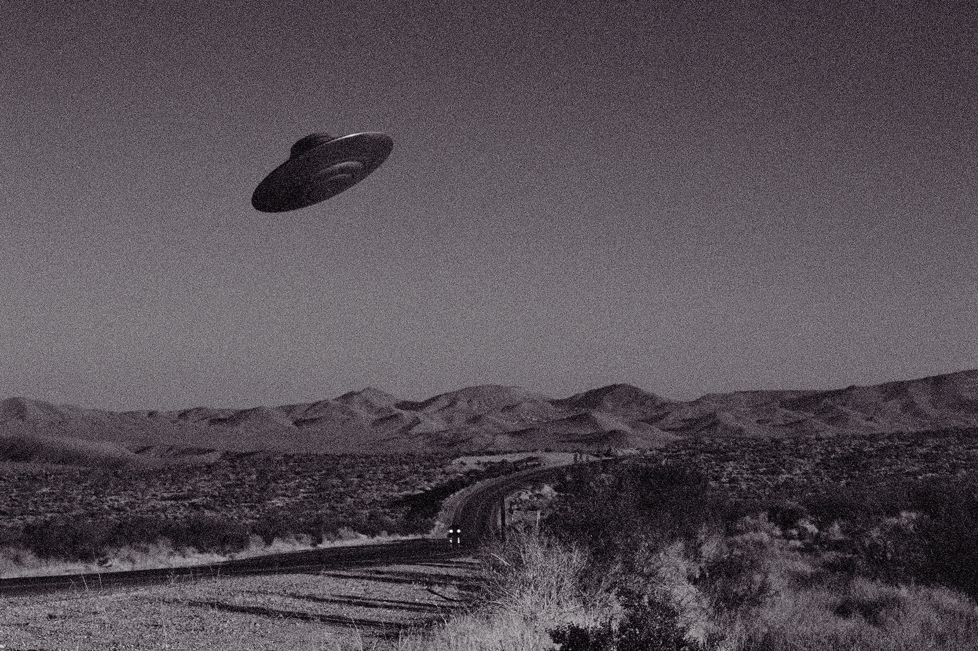 A black-and-white image of a flying saucer-shaped UFO suspended in the air above an arid, hilly landscape.