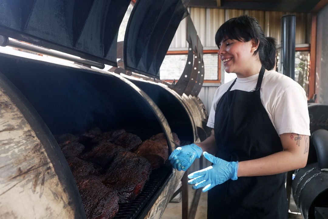Smiling woman wearing an apron stands next to a metal smoker.