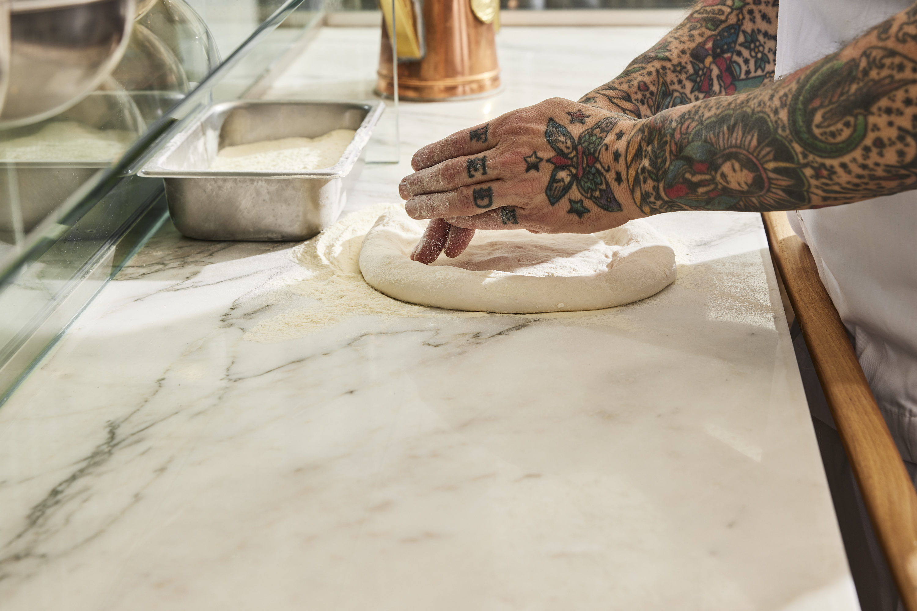 Tattooed hands massage a ball of pizza dough on a marbled counter.