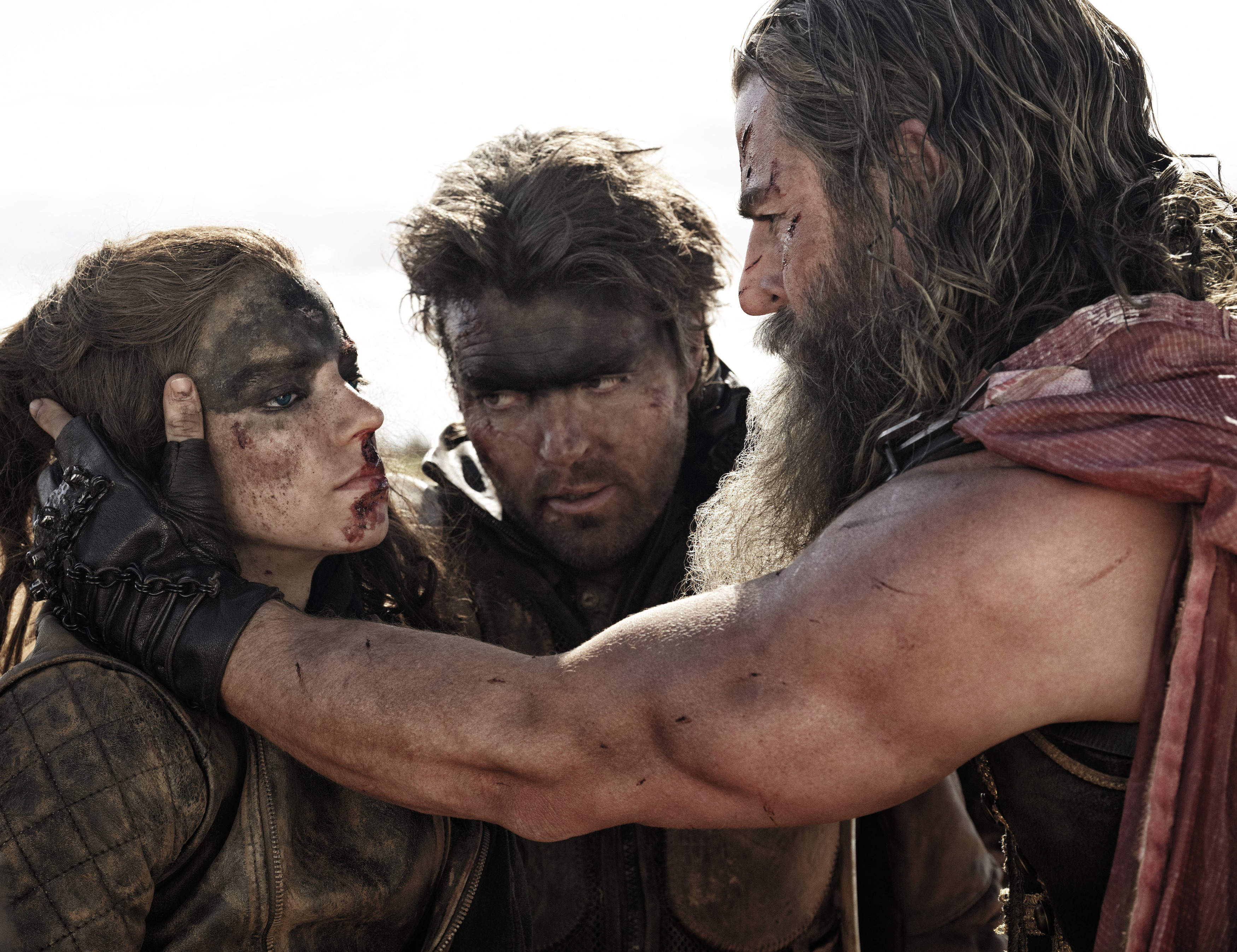 Furiosa (Anya Taylor-Joy), Praetorian Jack (Tom Burke), and Dementus (Chris Hemsworth), three extremely filthy Wasteland-dwellers with greasepaint-smeared faces, huddle close together, with Dementus holding Furiosa’s proud face in his hands, in George Miller’s Furiosa
