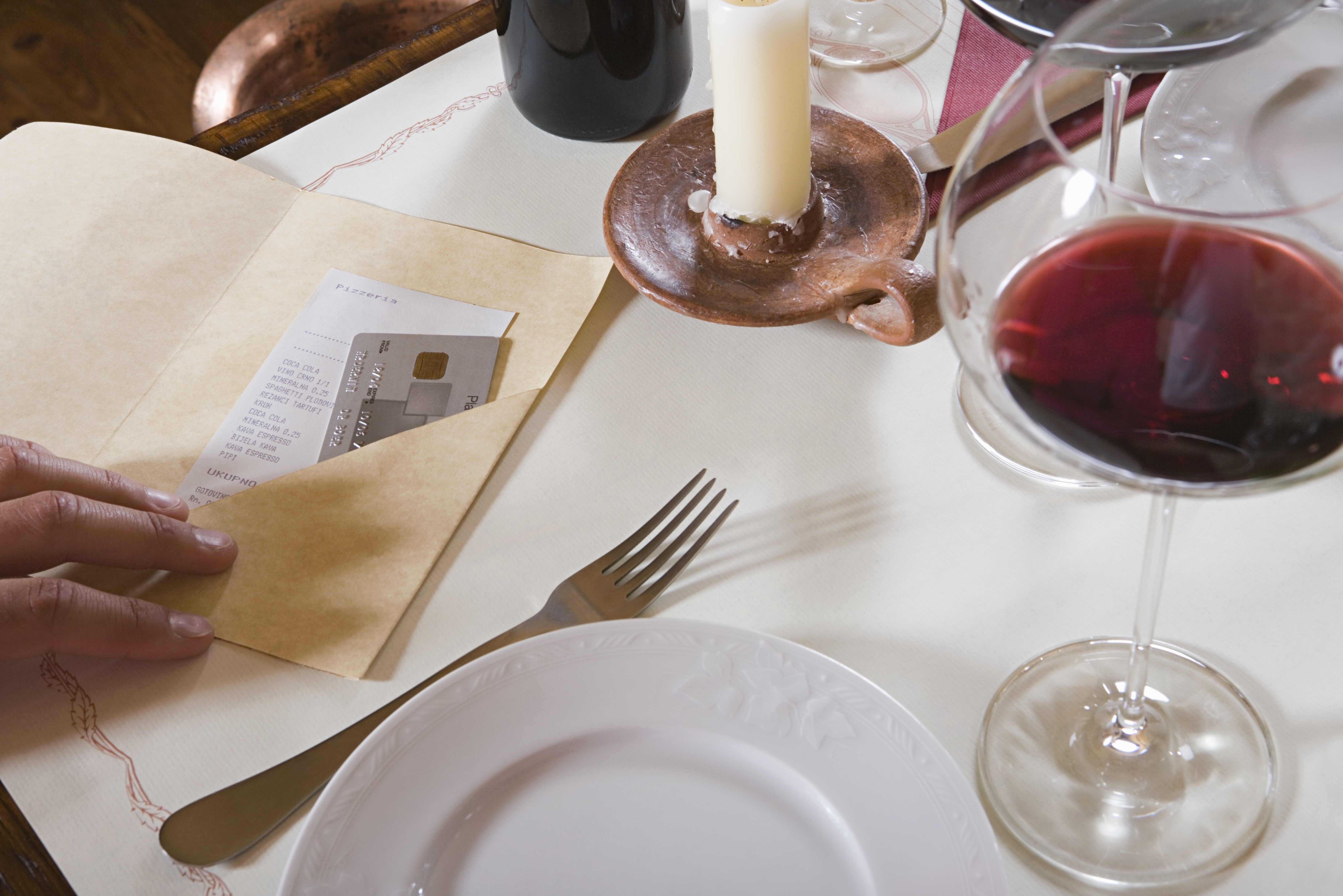 A white table set with a white plate, a fork, a glass of red wine, a candle, and an a manilla bill fold with a restaurant check in it; to the left of the frame, a hand is placing a credit card into the manila folder. 