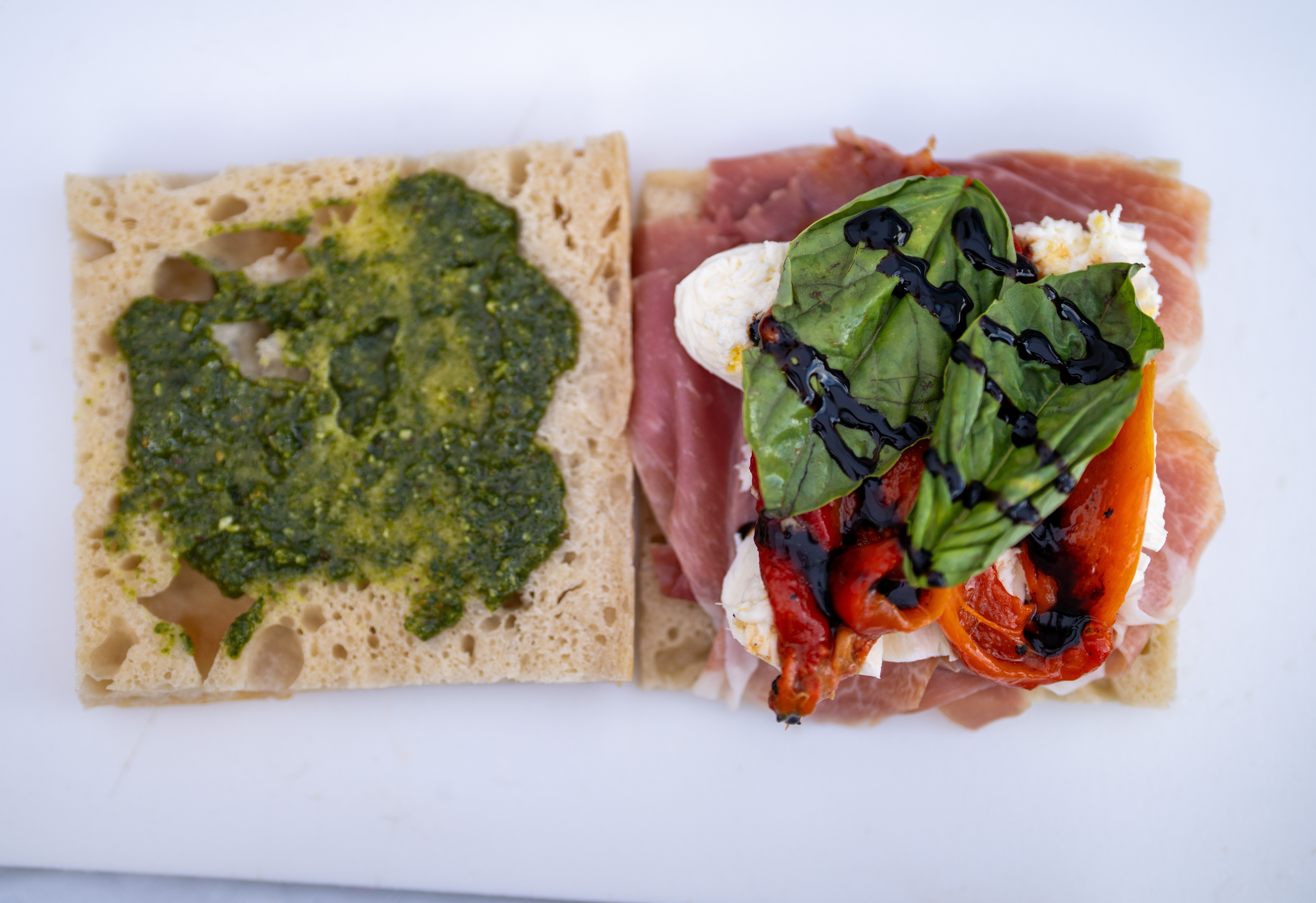 Overhead view of an open-faced sandwich; one side is slathered with pesto and the other layered with prosciutto, fresh mozzarella, red peppers, basil, and a balsamic glaze.